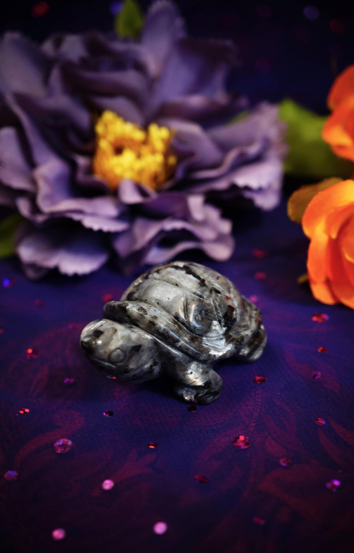 A small polished gray stone turtle figurine resting on a dark purple glittery surface, with large blurred purple and orange flowers in the background.