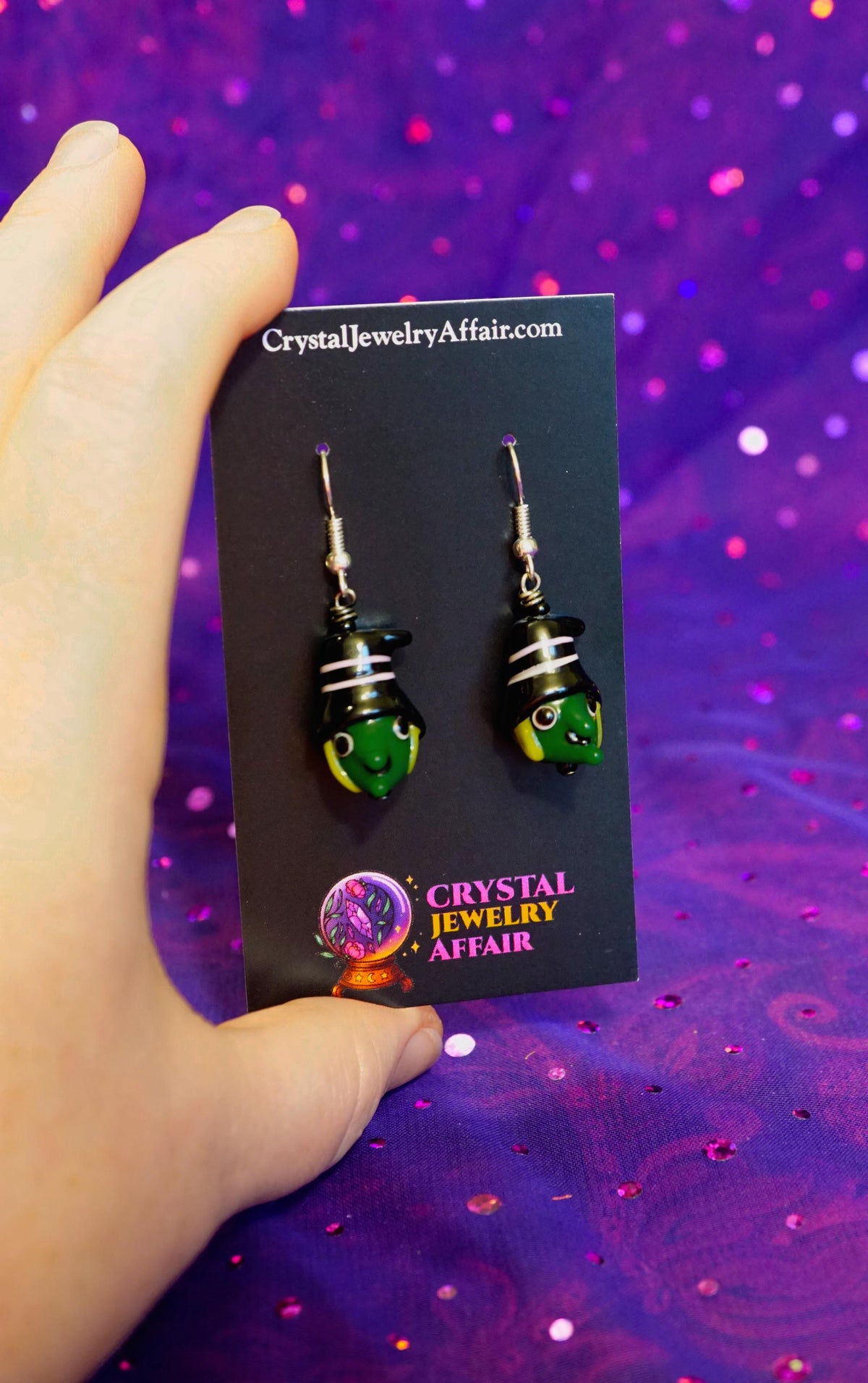 A hand holds a black display card featuring a pair of handmade glass dangle earrings. The earrings have green, black, and bright yellow witch heads.