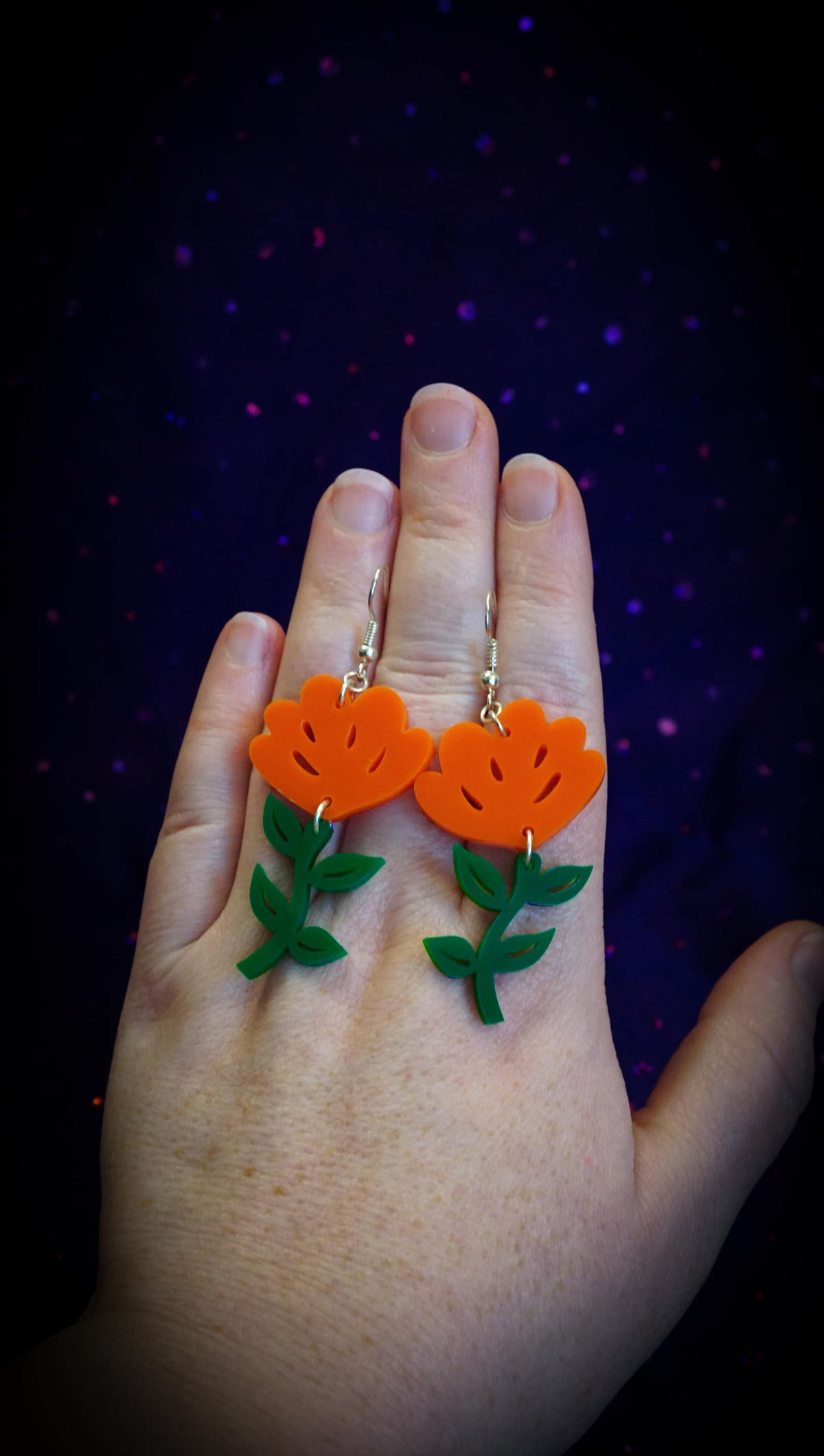 A hand wearing a pair of orange flower-shaped drop earrings with green leafy stems and gold hooks, set against a dark starry background.