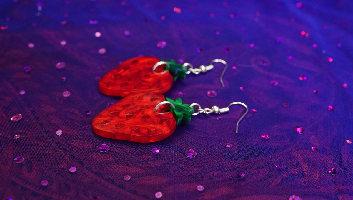 A pair of strawberry-shaped dangling earrings with translucent red bodies, green leafy tops, and silver wires, displayed on a purple glittery fabric background.