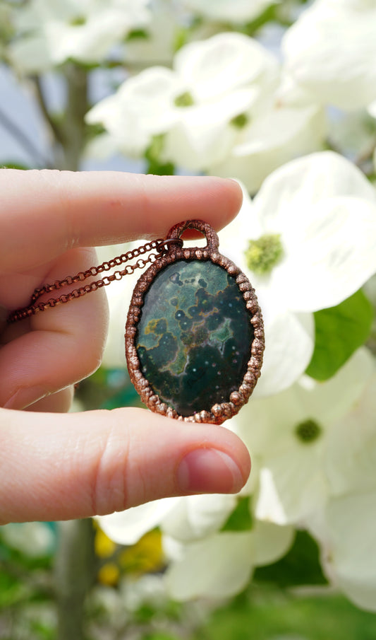 Hand holding a copper pendant with a green stone against a blurred floral background