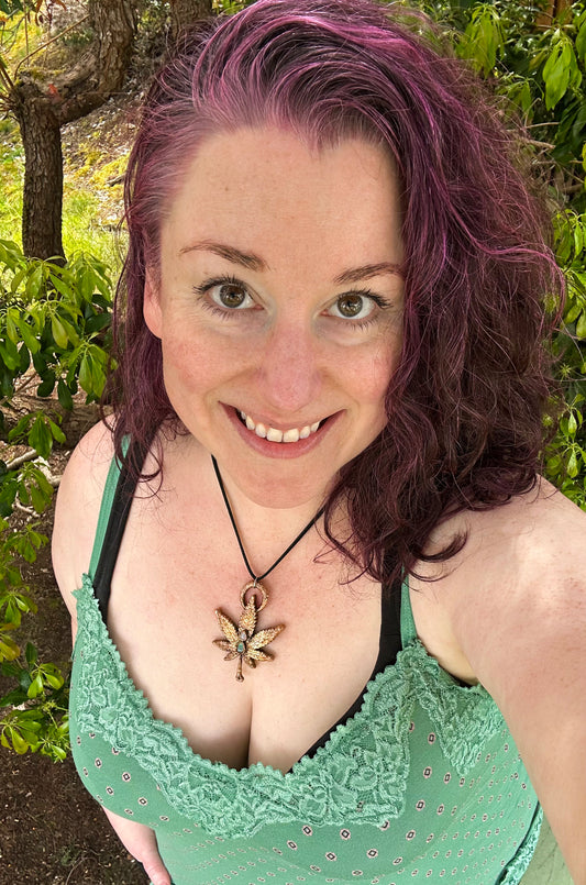 Woman with purple hair wearing a green top and weed necklace in a forest setting