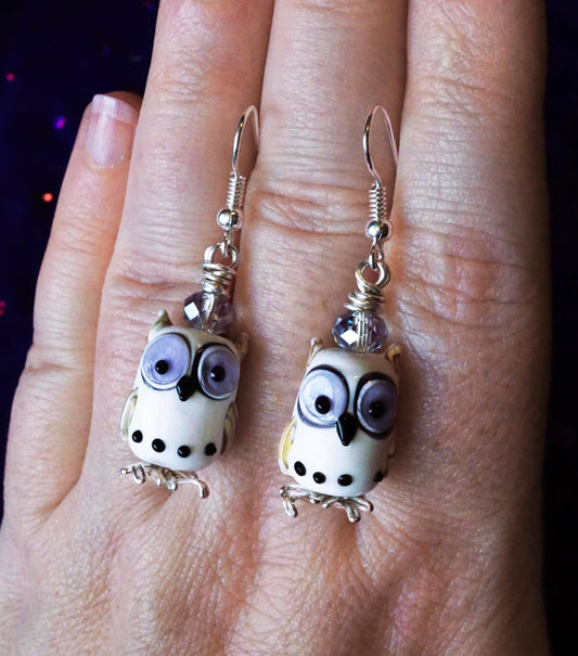 white Owl-shaped earrings on a hand against a dark background