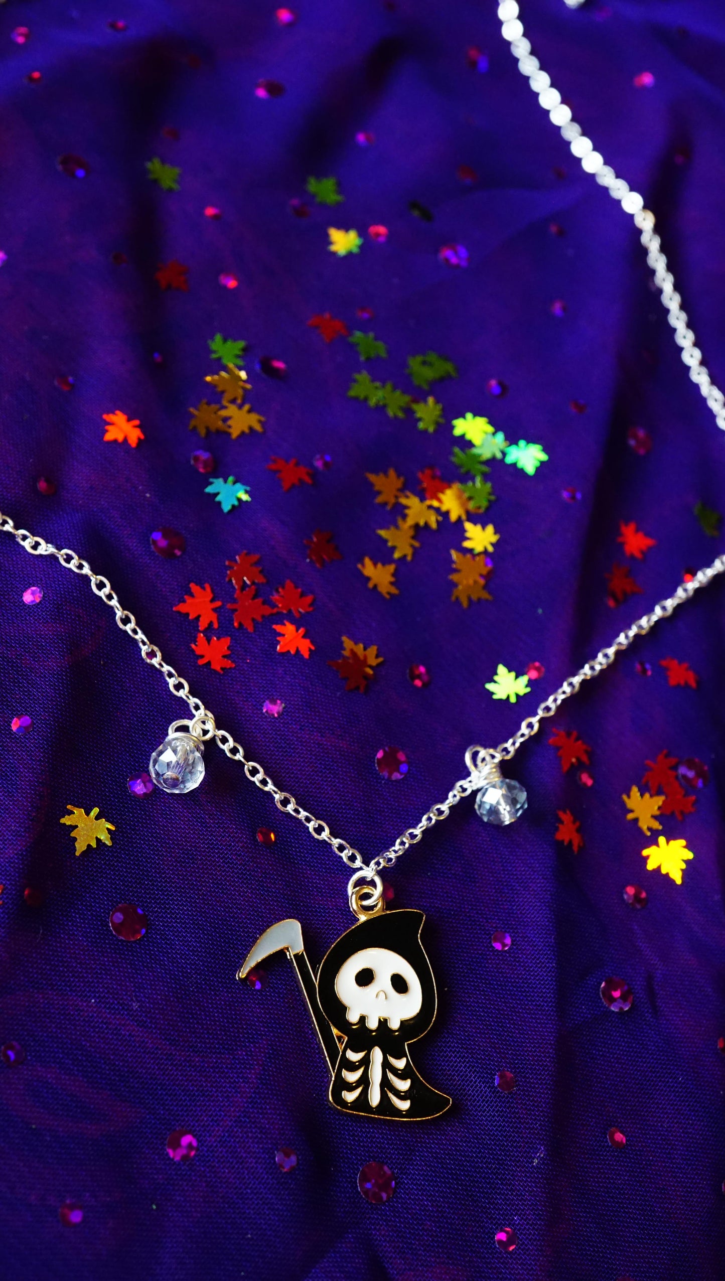 skeleton reaper charm necklace with small blue-grey beads dangling. silver toned chain. purple background with rainbow leaf confetti that flashes beautifully.