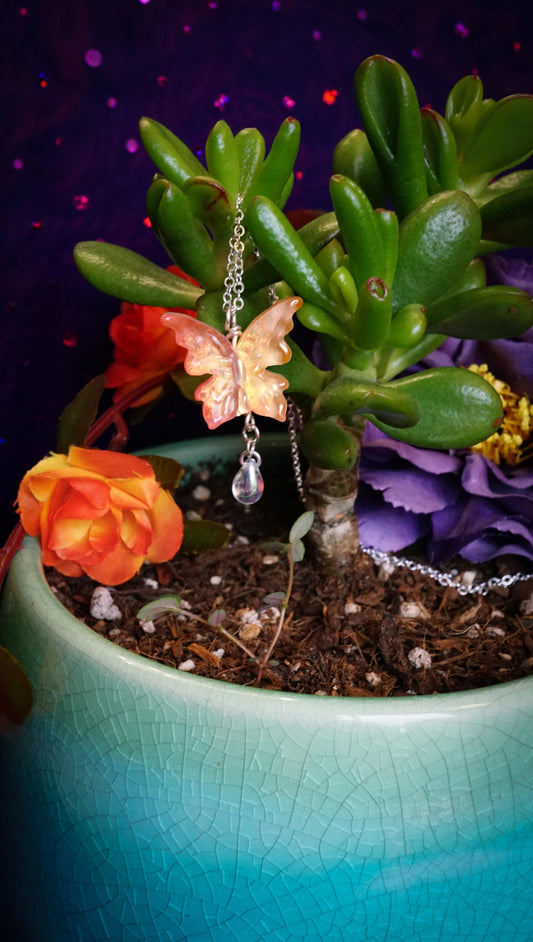 An orange and yellow, peachy butterfly-shaped drop necklace with a clear teardrop charm hangs from a succulent plant in a ceramic pot with orange and purple flowers against a dark purple background.