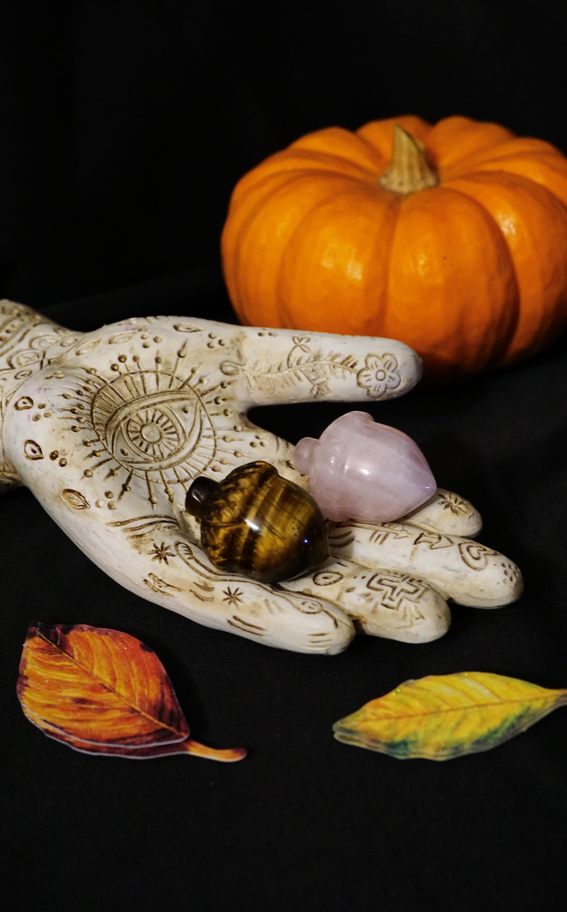 Decorative hand with henna designs holding pink and brown acorn crystal figurines, all surrounded by a pumpkin and leaves on a dark background