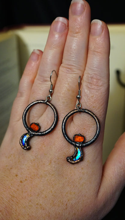 flashy crescent moon crystals below orange jack o' lanterns, set in a sturdy copper setting. placed on hand for size reference. deep ombre copper effect.