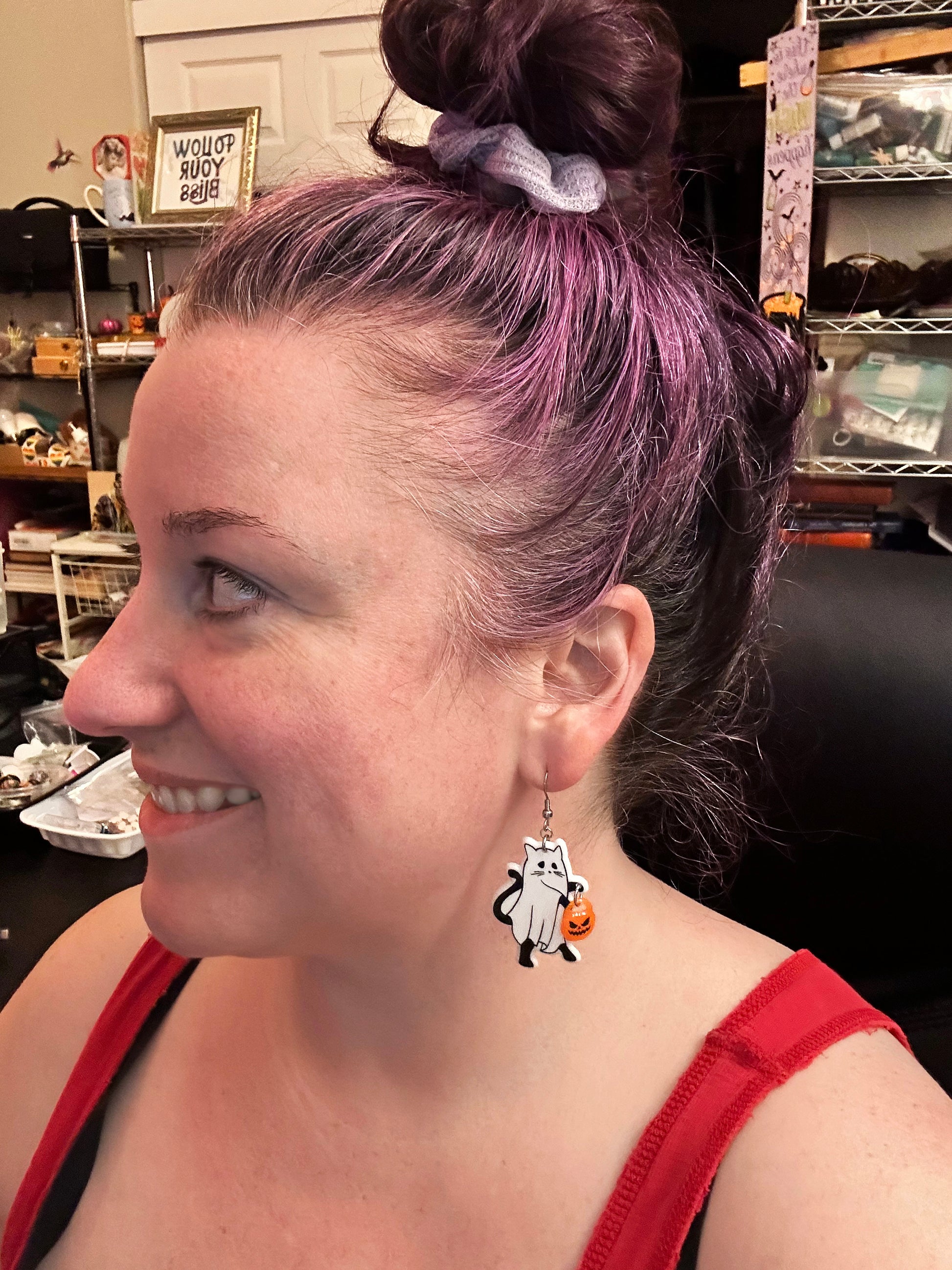 Person with purple hair styled in a bun, wearing a red top and halloween ghost cat-themed earrings.