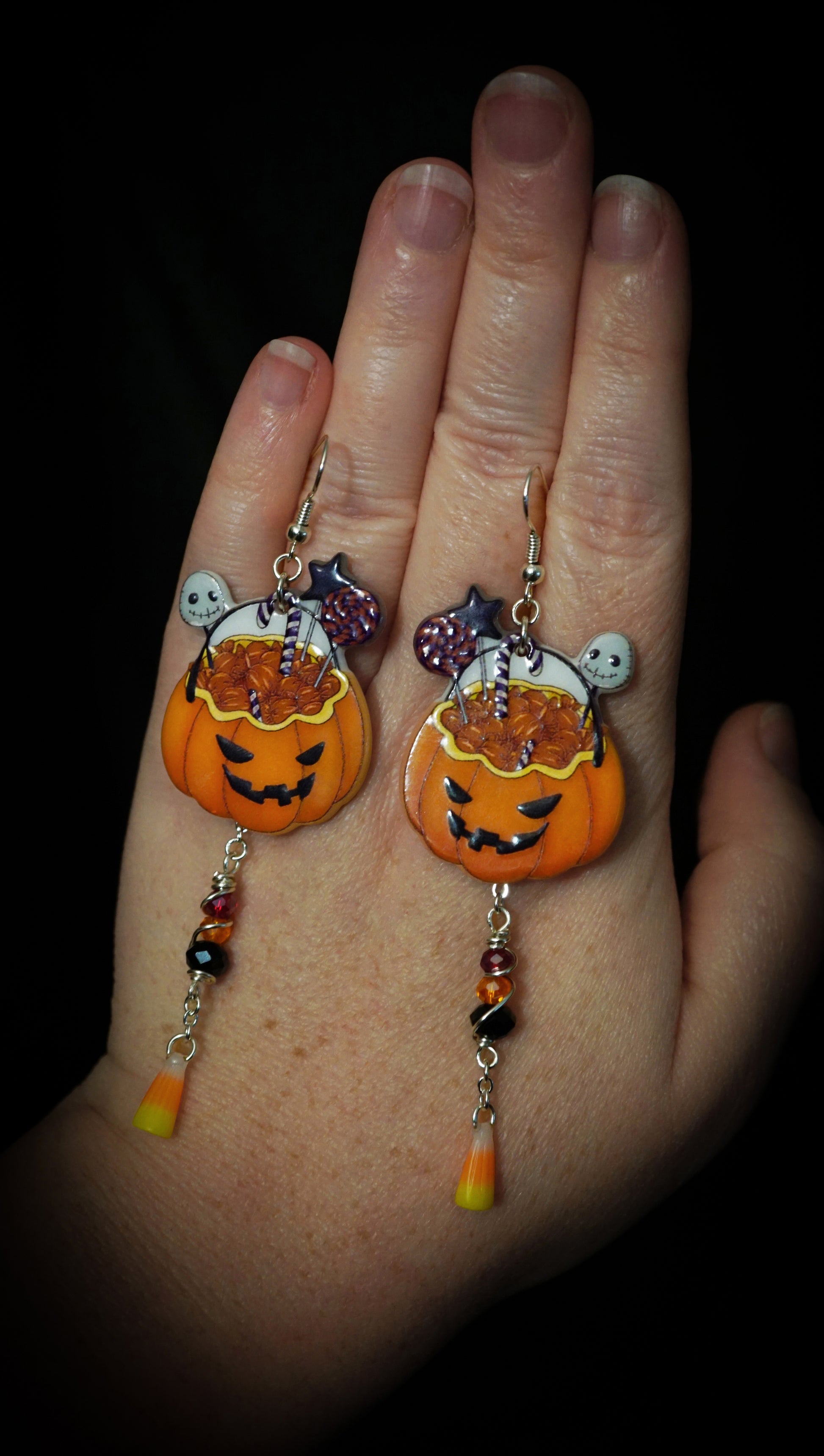 orange and black Pumpkin-shaped earrings with wire wrapped beads in orange, black and red tones. miniature candy corns hang below beads. on hand. black background.