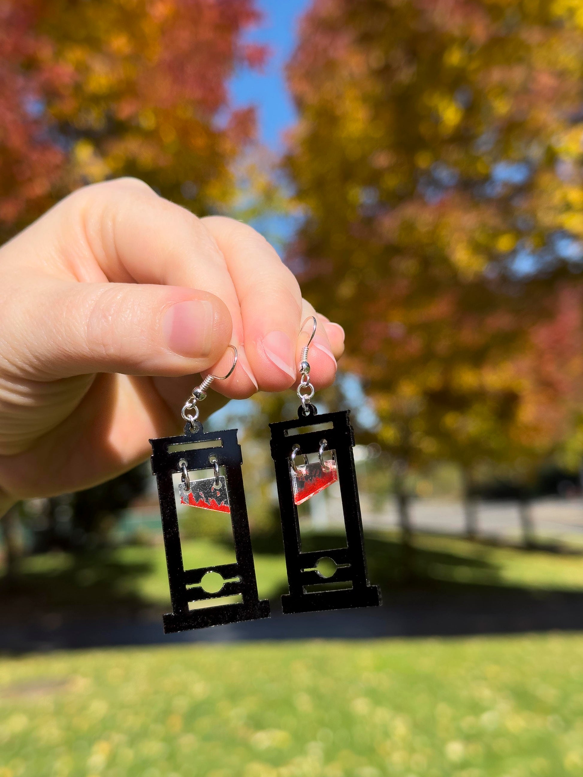 hand holding black guillotine earrings with red bloody blades that dangle. fall tree background
