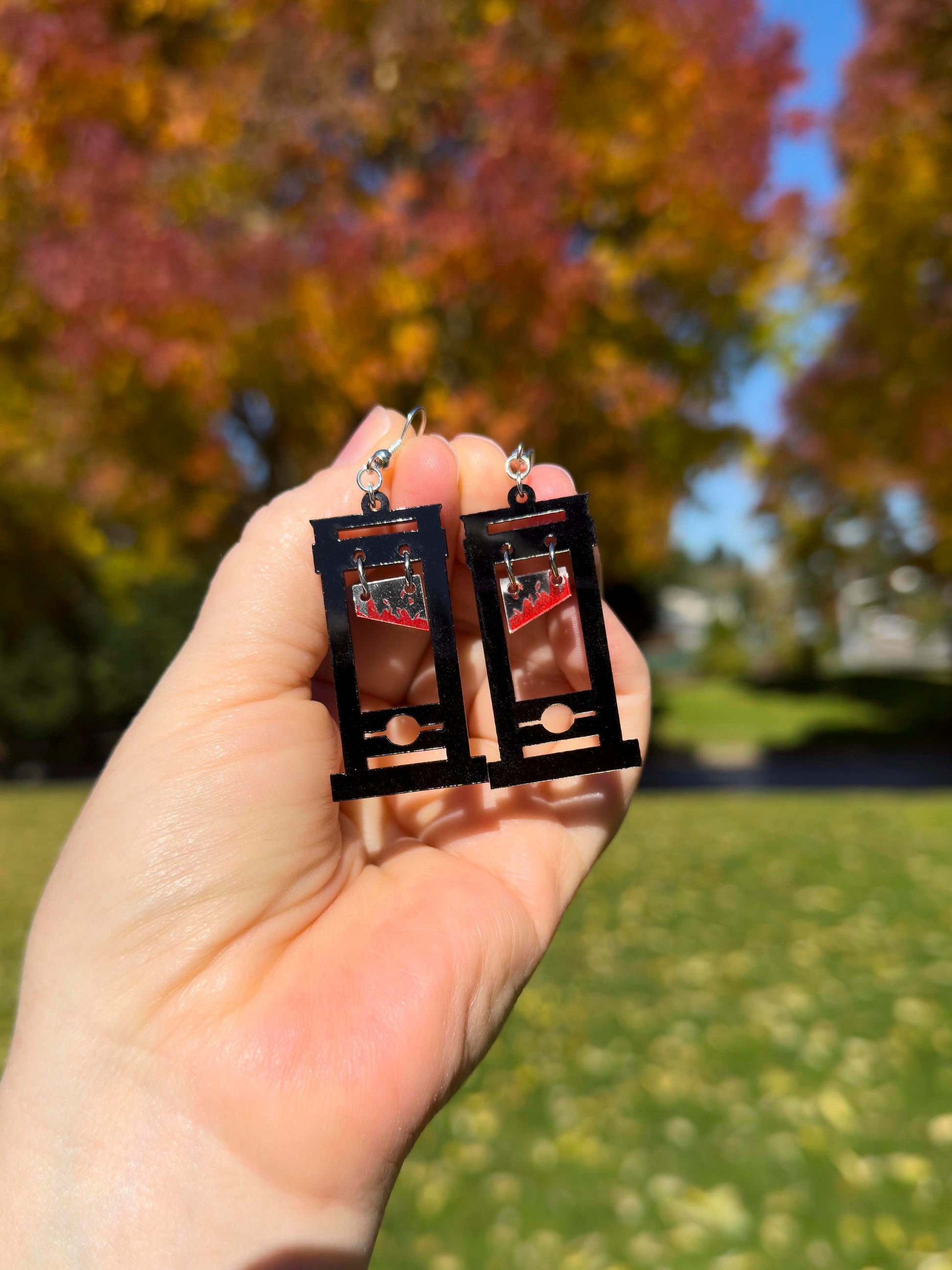 hand holding black guillotine earrings with red bloody blades that dangle towards the top. don't lose your head! orange, yellow and red fall foliage and grass in background