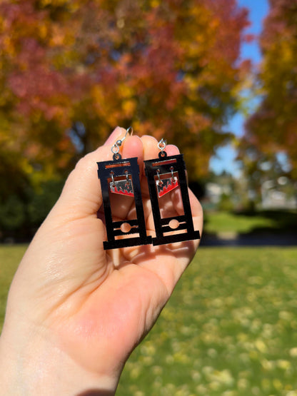 hand holding black guillotine earrings with red bloody blades that dangle towards the top. don't lose your head! orange, yellow and red fall foliage and grass in background