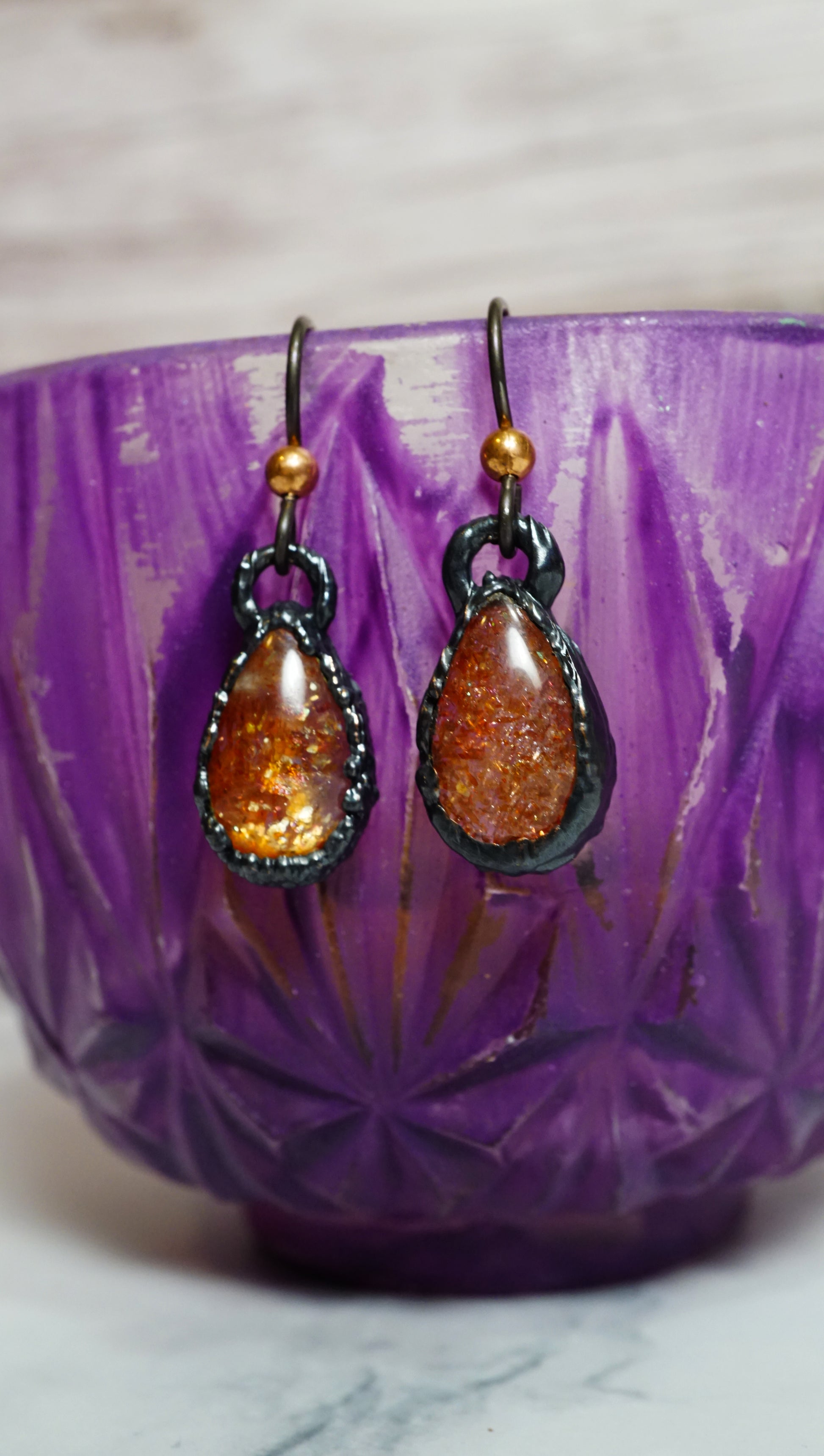 orange crystal earrings in teardrop shape. dainty black copper settings hanging on purple teacup