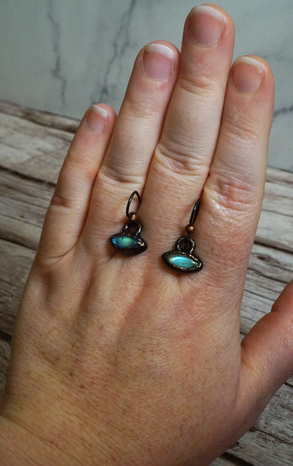 Hand holding a set of dainty blue and green crystal earrings with dark brown/copper settings. shaped like spaceships or eyes. fake wood and marble background