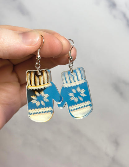 Mitten-shaped earrings with blue and white design held by a hand against a neutral background