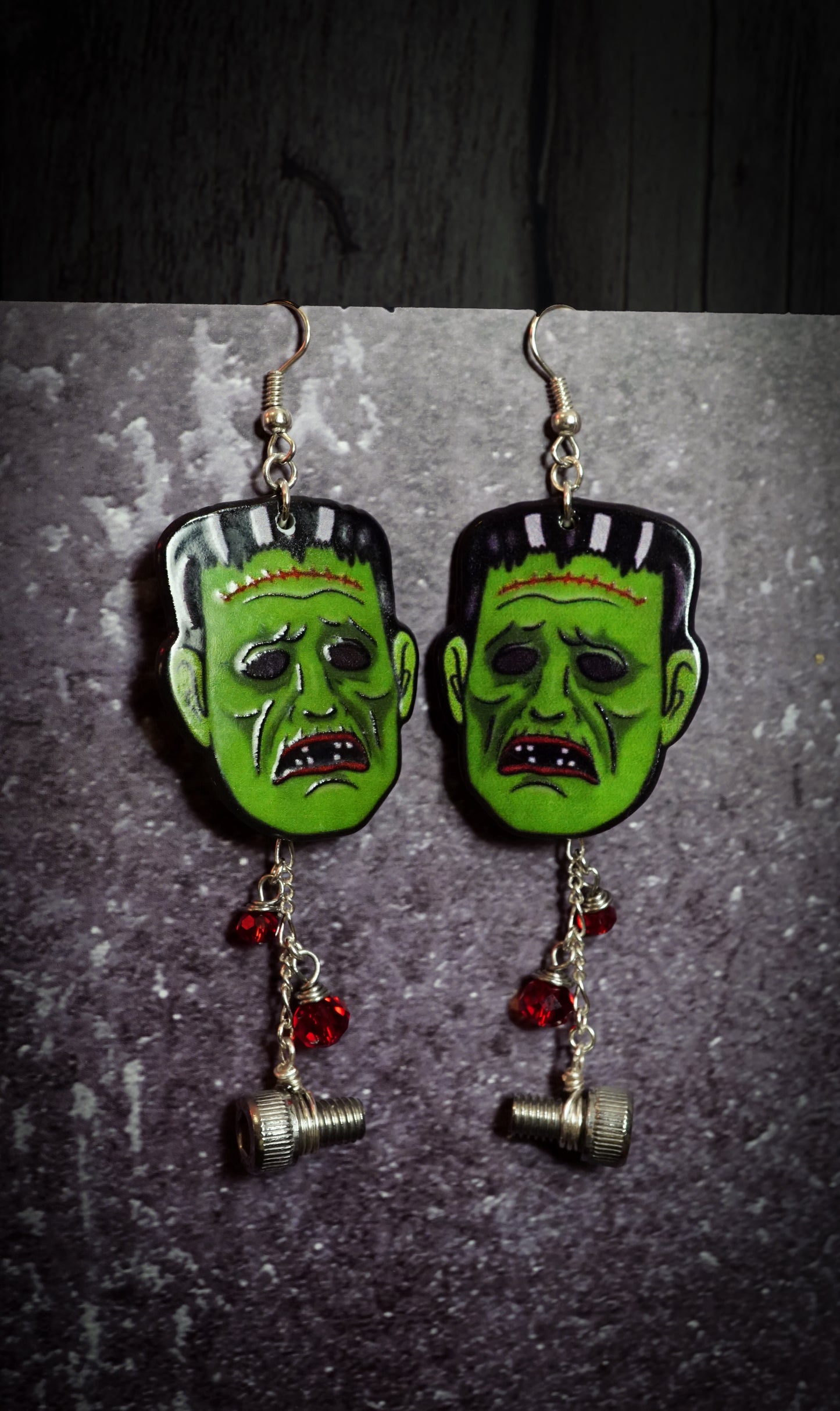 Pair of earrings with green monster faces and red beads on a dark background