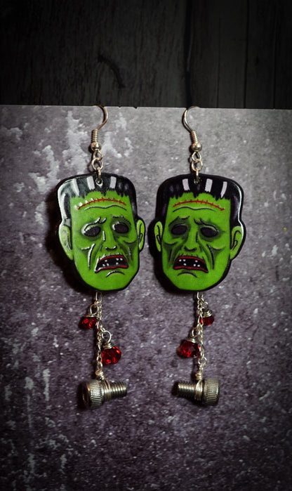 Pair of earrings with green monster faces and red beads on a dark background