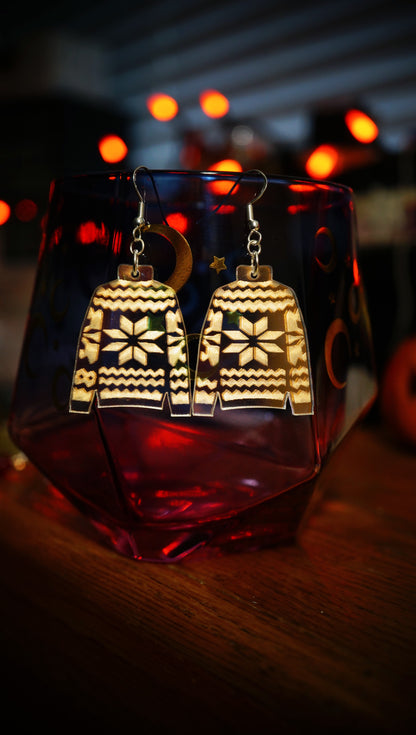 Pair of earrings shaped like sweater designs on a glass with blurred lights in the background