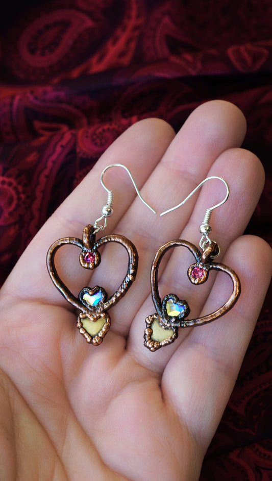 Heart-shaped hoop earrings with colorful stones held in a hand against a red fabric background