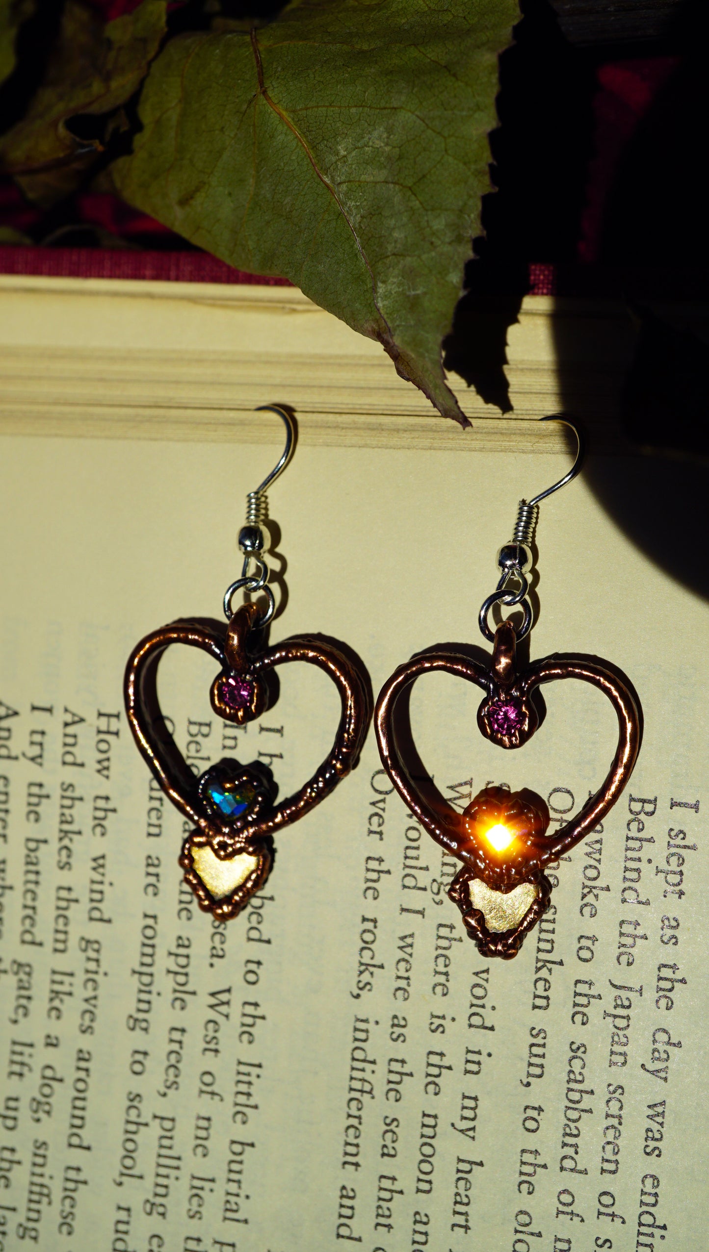 heart shaped dangles with pink rhinestones, brass hearts, and aurora borealis heart stones. hearts are sparkling with a sunburst from the sun. against an old book with a green leaf.
