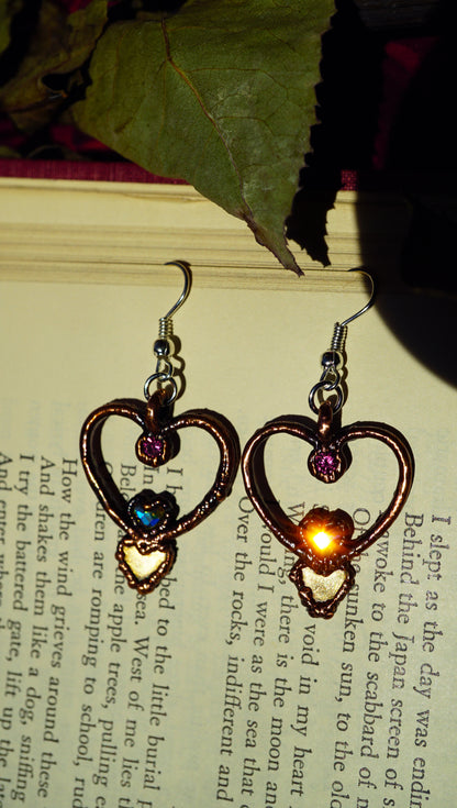 heart shaped dangles with pink rhinestones, brass hearts, and aurora borealis heart stones. hearts are sparkling with a sunburst from the sun. against an old book with a green leaf.