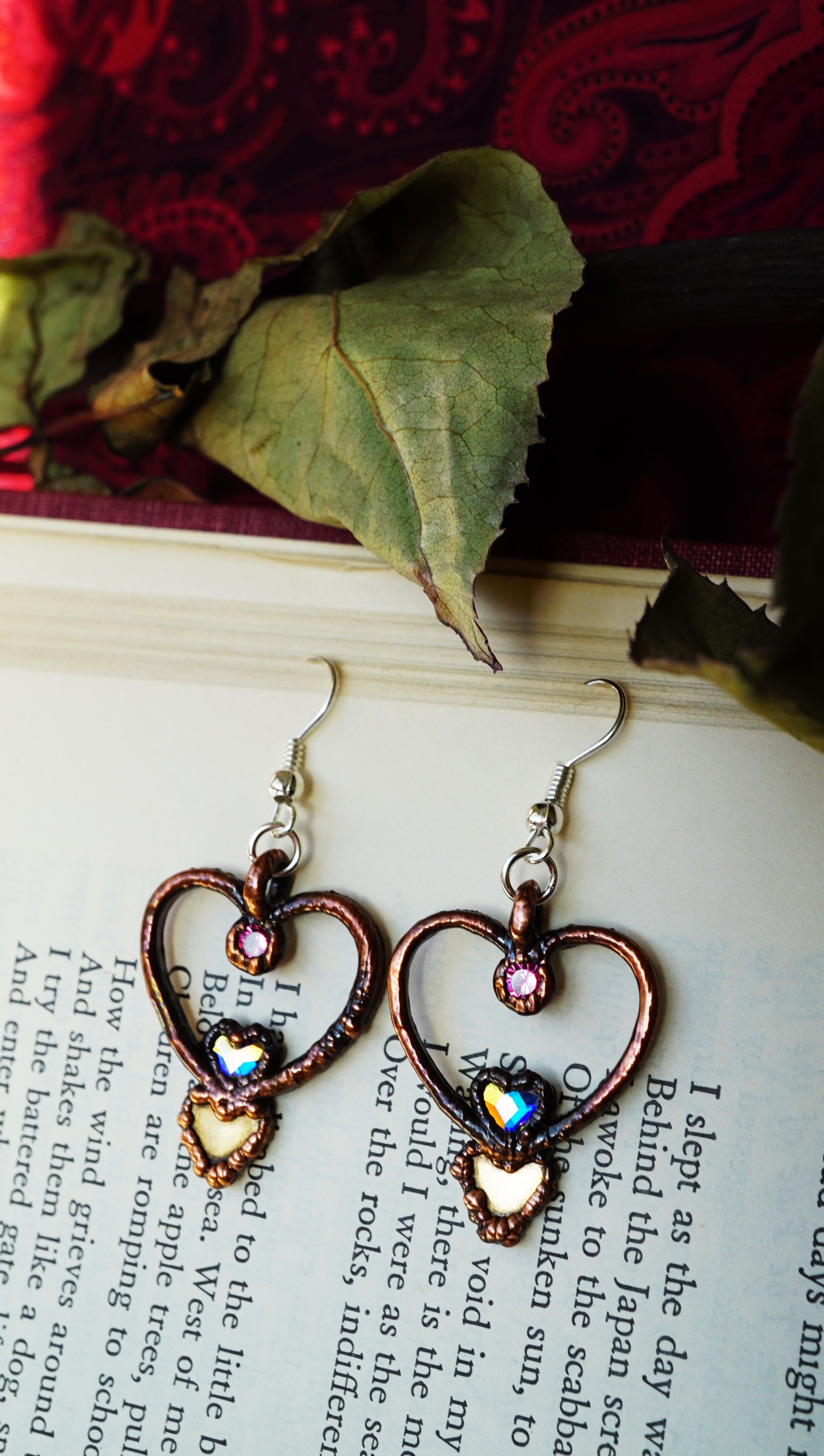Heart-shaped earrings with colorful stones on a book page with red fabric and leaves in the background.