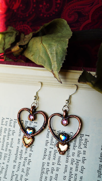 Heart-shaped earrings with colorful stones on a book page with red fabric and leaves in the background.