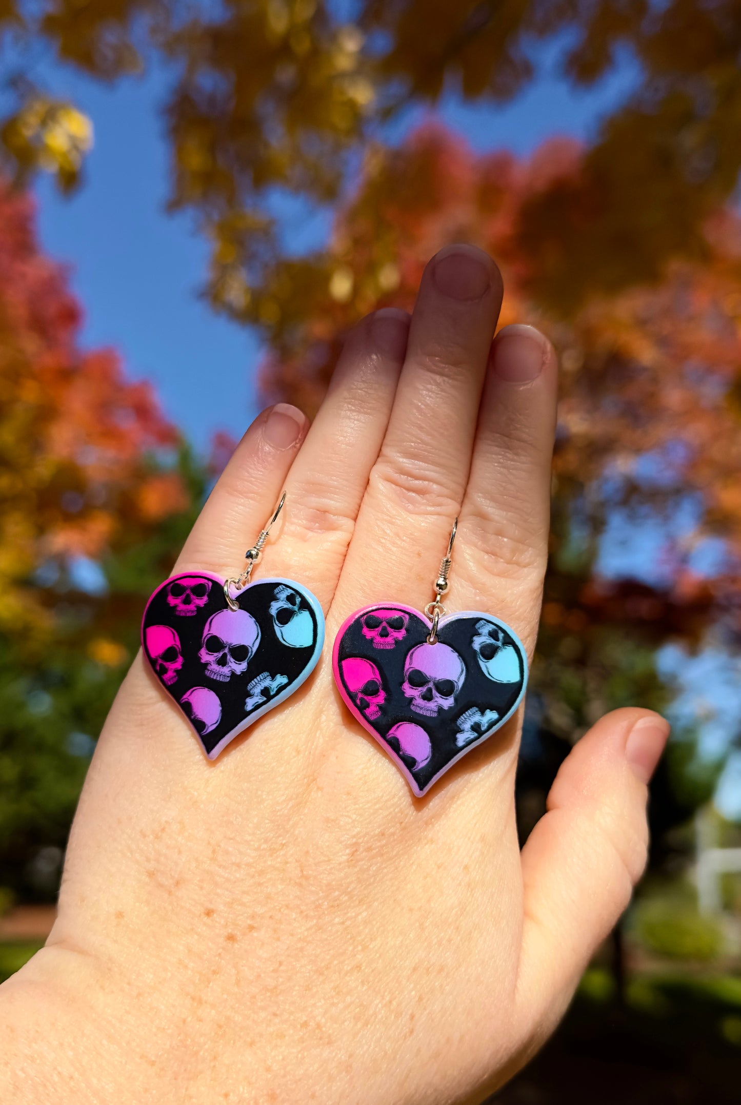 hand holding dangle heart earrings. hearts are black with vibrant pink, purple, light blue skulls. orange and red Fall trees in background