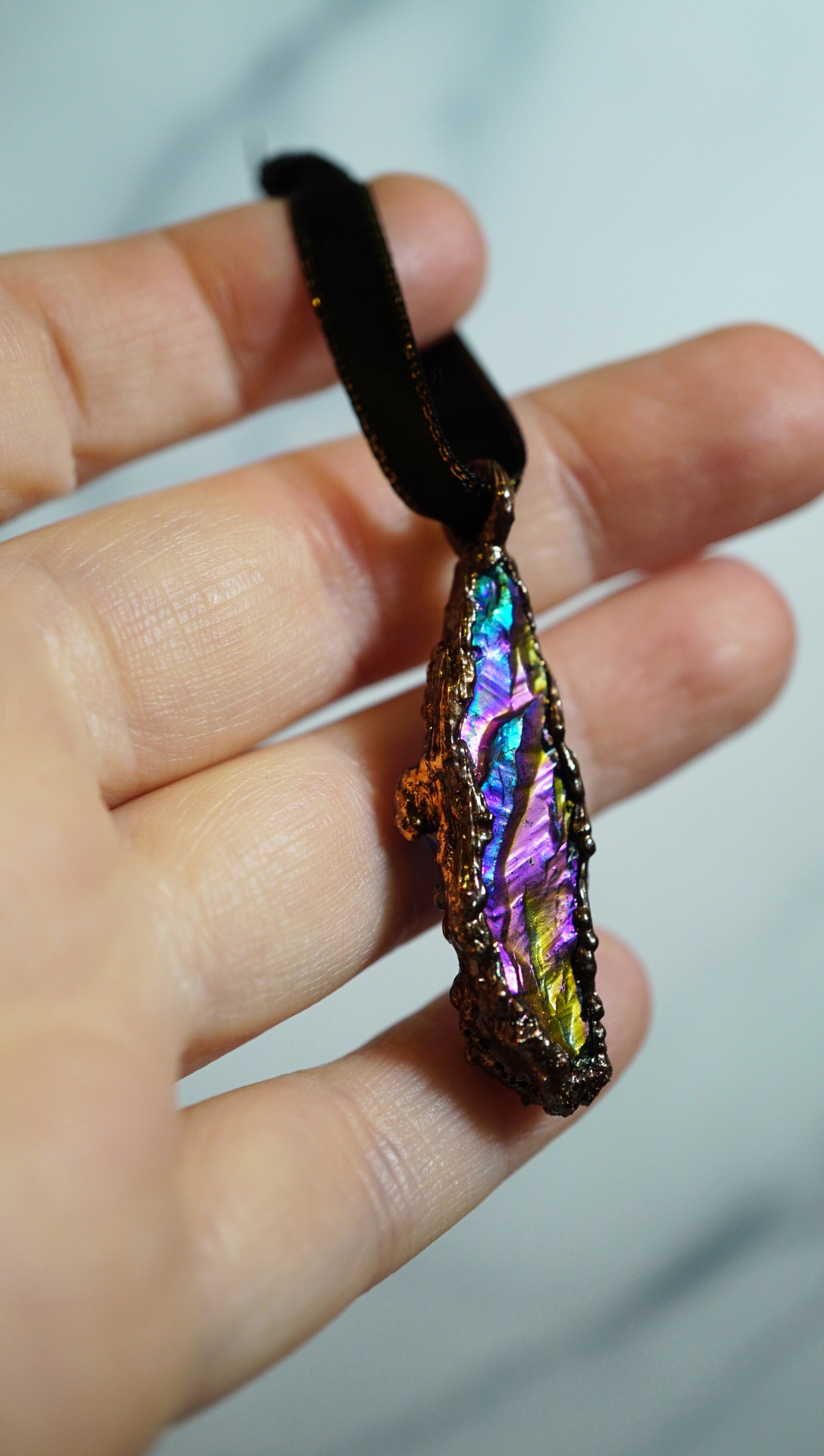 rainbow colored crystal ornament in copper setting, black ribbon. in hand against blurry grey background