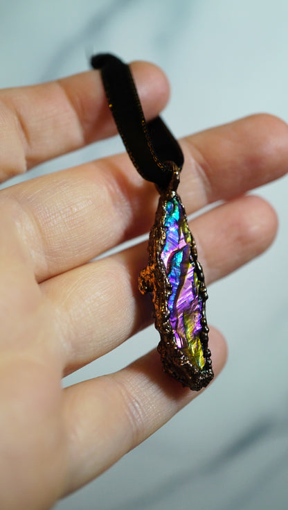 rainbow colored crystal ornament in copper setting, black ribbon. in hand against blurry grey background
