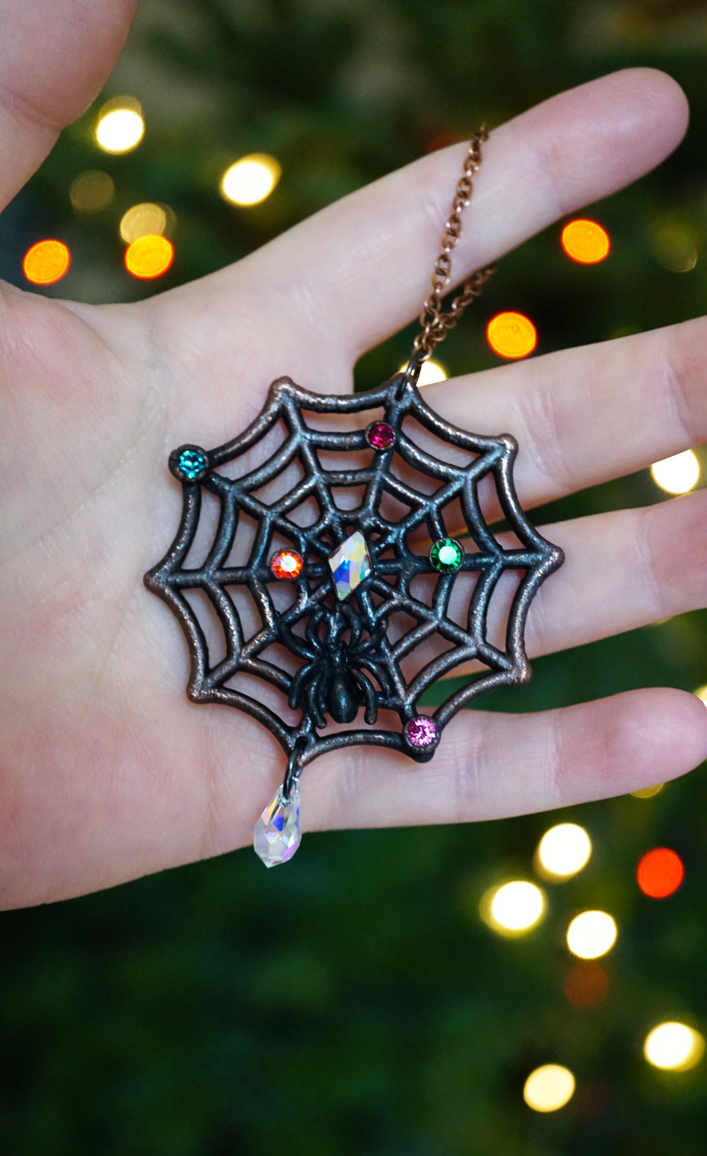 dark copper spiderweb ornament with small christmas light rhinestones, black spider, in hand against a lit christmas tree