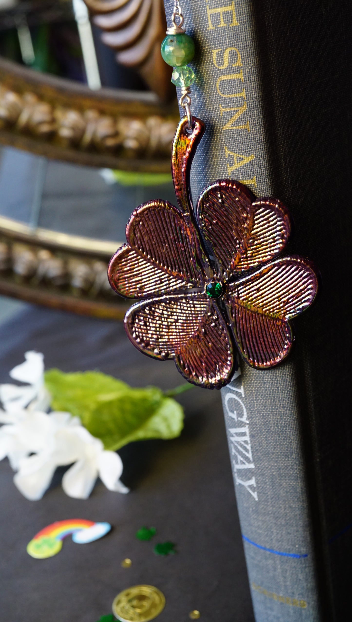 shamrock bookmark on old book, green crystal details, background has gold coins, shamrocks and white flowers by a mirror