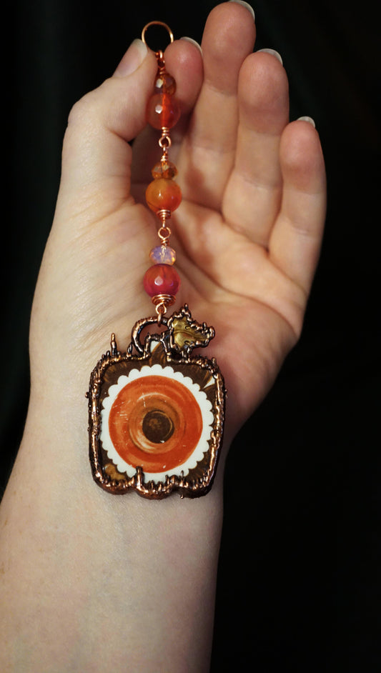 A hand holding a wall hanging with pink and orange agate beads with copper wire, and a pumpkin portal looking broken china piece, electroformed in spiky textured copper. Small brass leaf. black background behind hand.