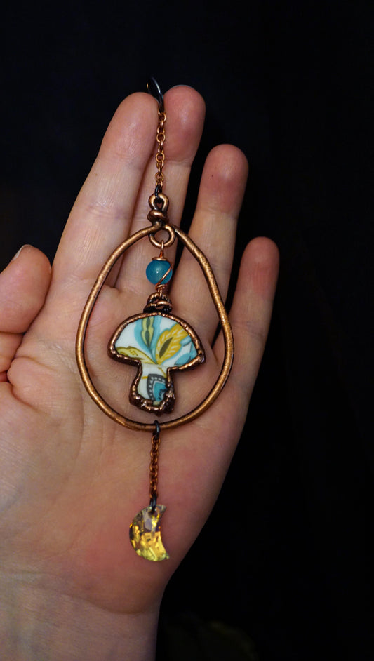 broken china mushroom wall hanging or suncatcher with moon dangle and blue bead. held against hand and black backround