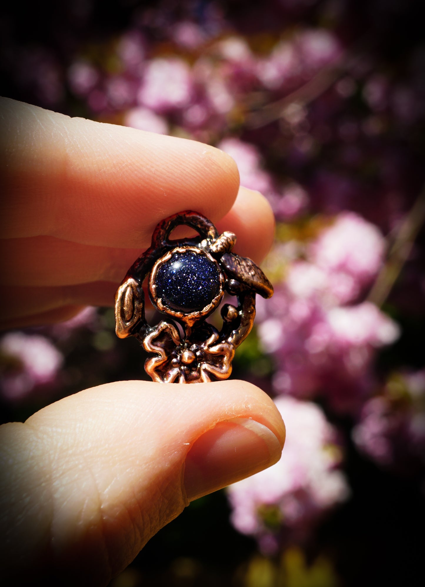 Hand holding a decorative pendant with a blue stone against a blurred floral background
