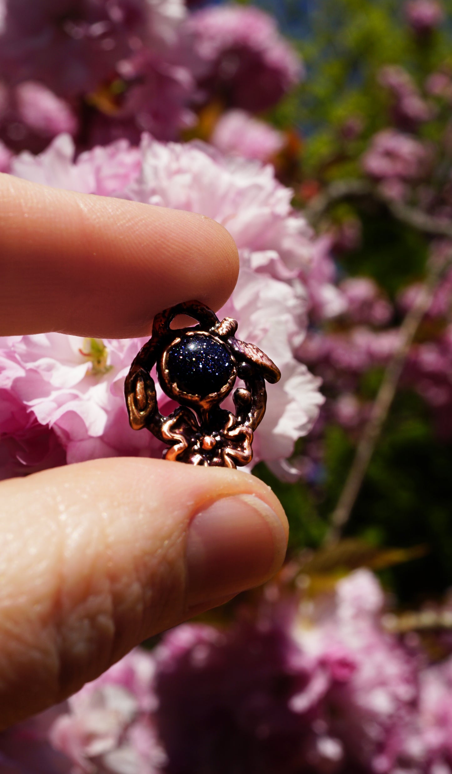 Person holding a small bronze pendant with a blue stone against a pink flower background