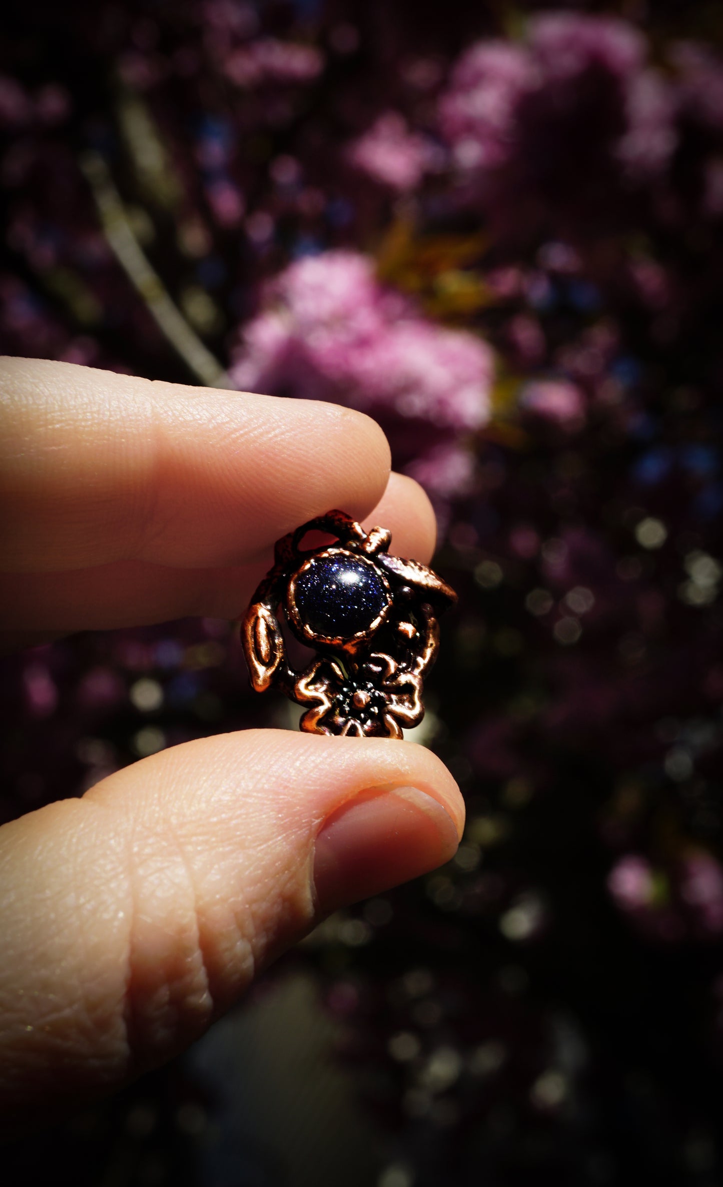 hand holding pendant for scale. dark blue sparkly stone in a copper cherry blossom setting, background is blurred pink cherry and plum blossoms