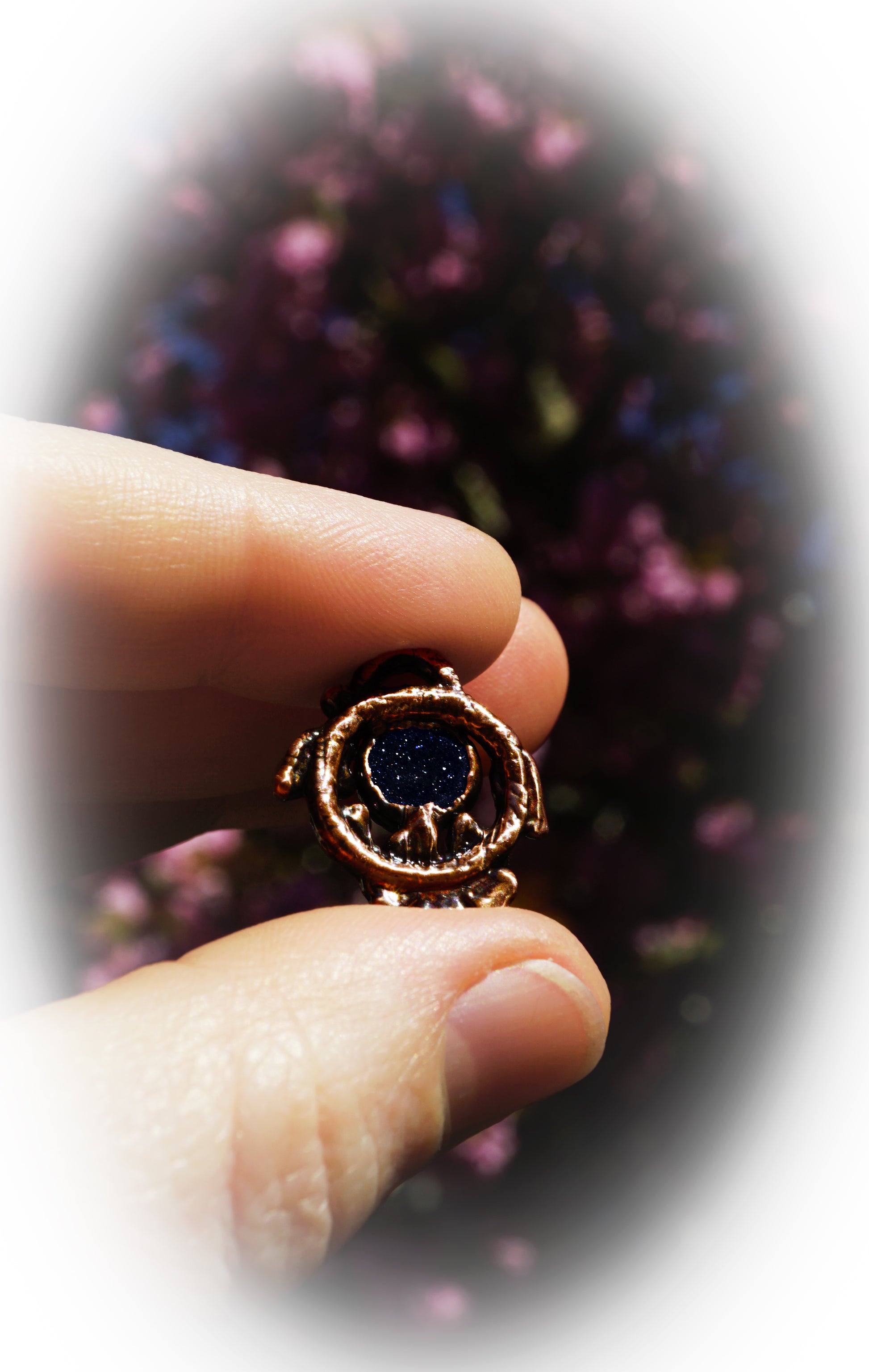 hand holding a copper pendant with dark blue stone, against pink cherry blossom background