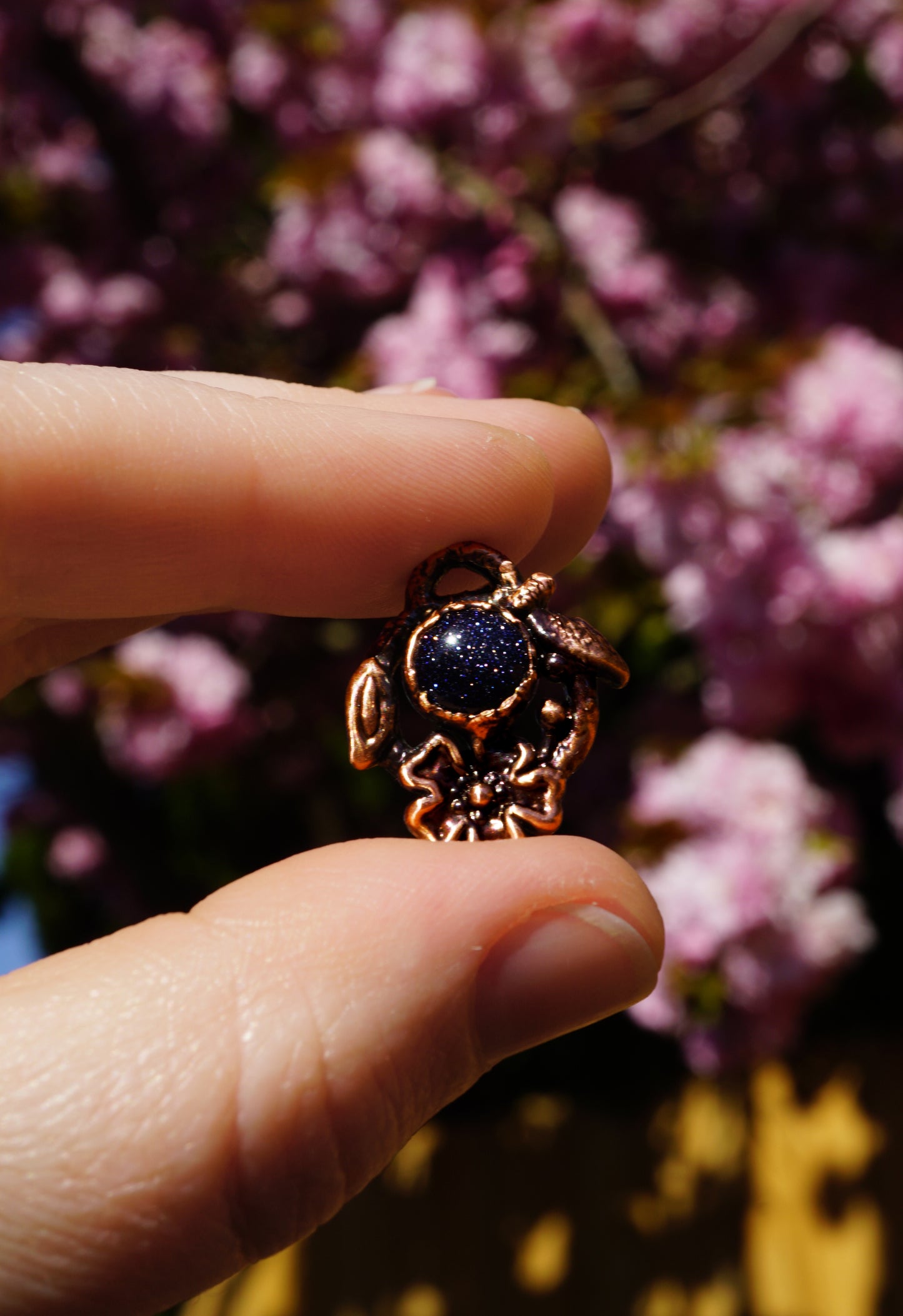 Hand holding a small blue stone and dark copper pendant against a blurred floral background