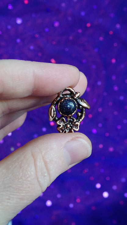 dark blue goldstone in dark copper floral setting, between fingers, purple fabric background