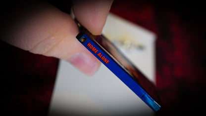 side of blue mini Home Alone DVD case charm, with red text, between fingers for size, red background