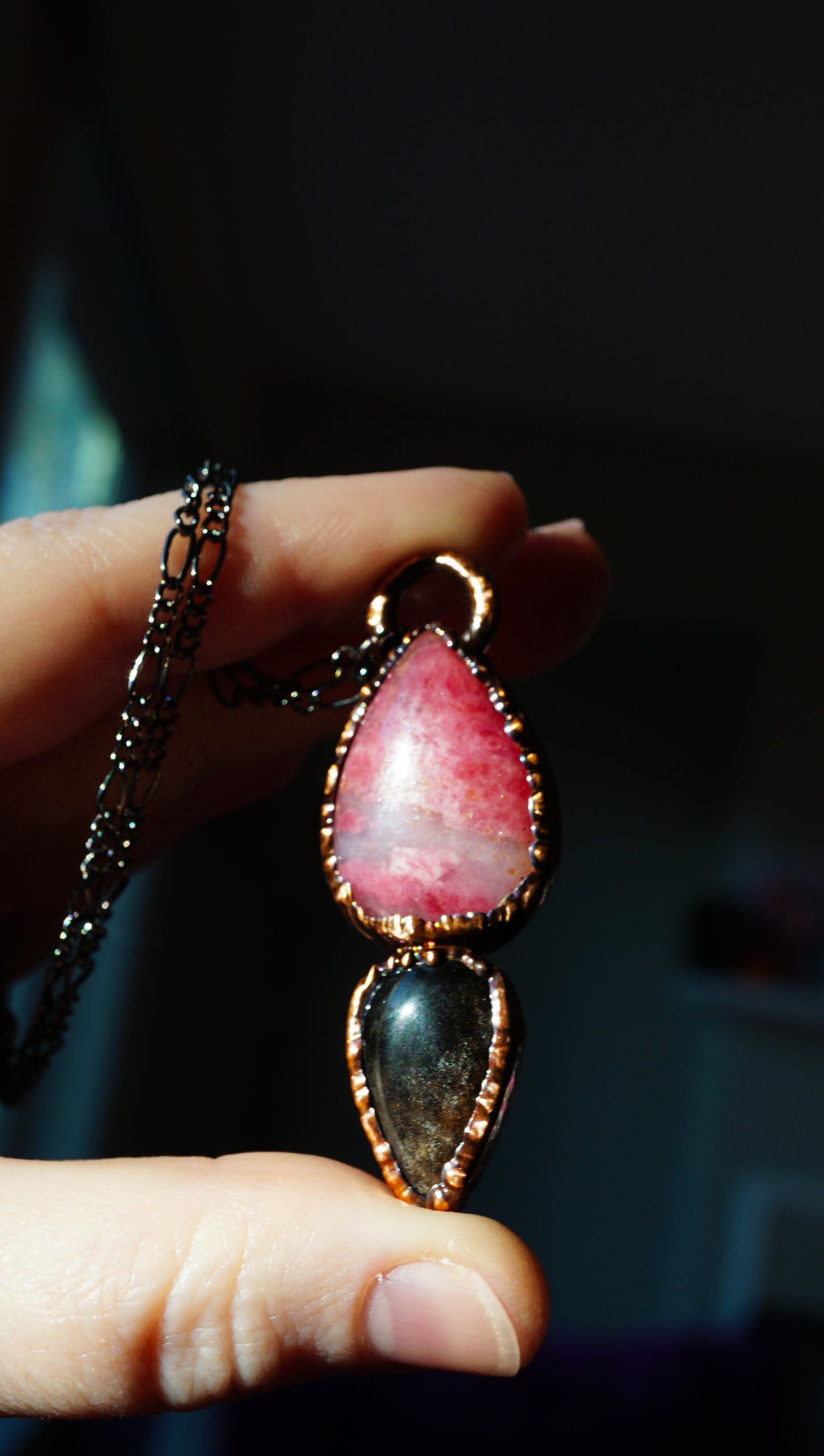 Hand holding a necklace with a pink and black pendant against a dark background