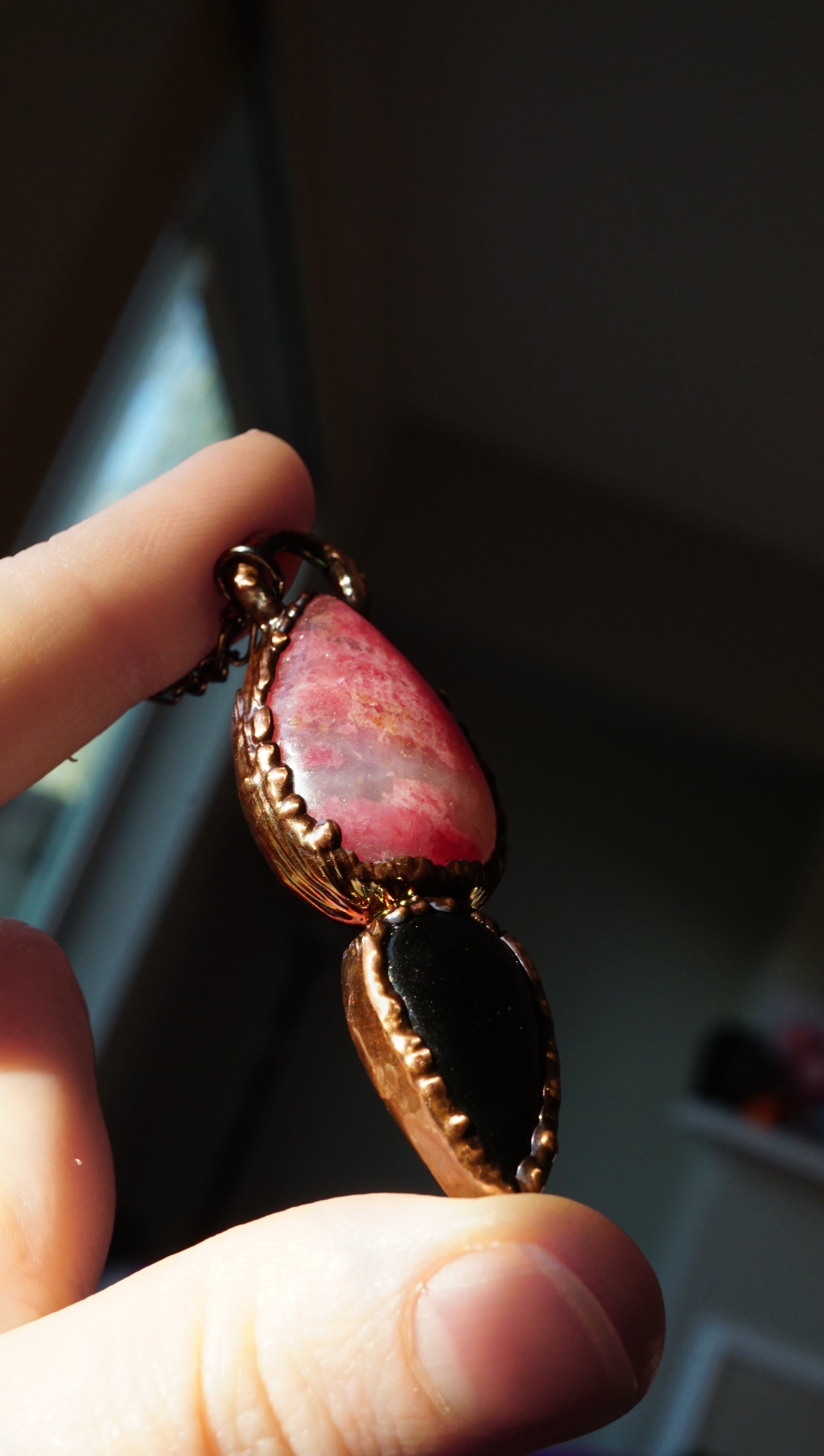 side view of pink rhodonite and obsidian necklace. textured copper around the stones. held in hand against dark background. 