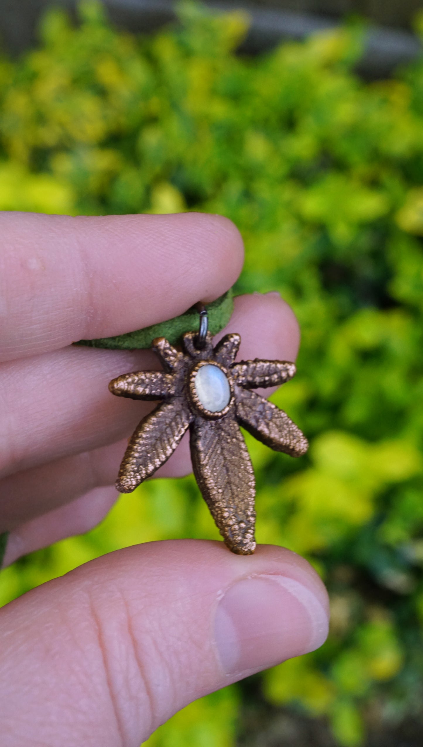 Bronze weed leaf shaped pendant with a central gemstone held between fingers against a blurred green background