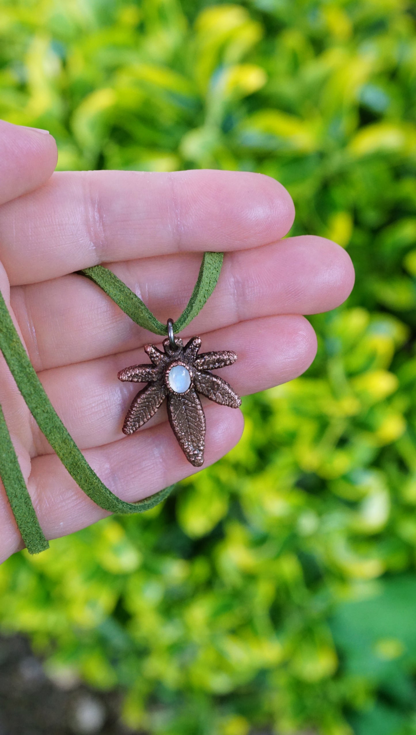 hand holding copper weed leaf pendant with white moonstone center, green vegan suede, against bright green bush