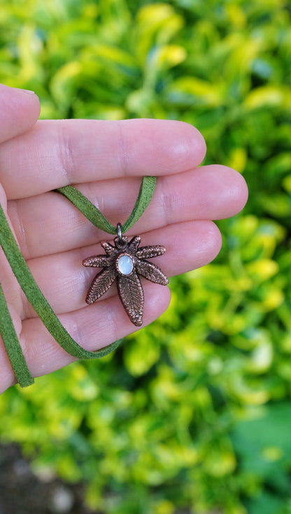 hand holding copper weed leaf pendant with white moonstone center, green vegan suede, against bright green bush