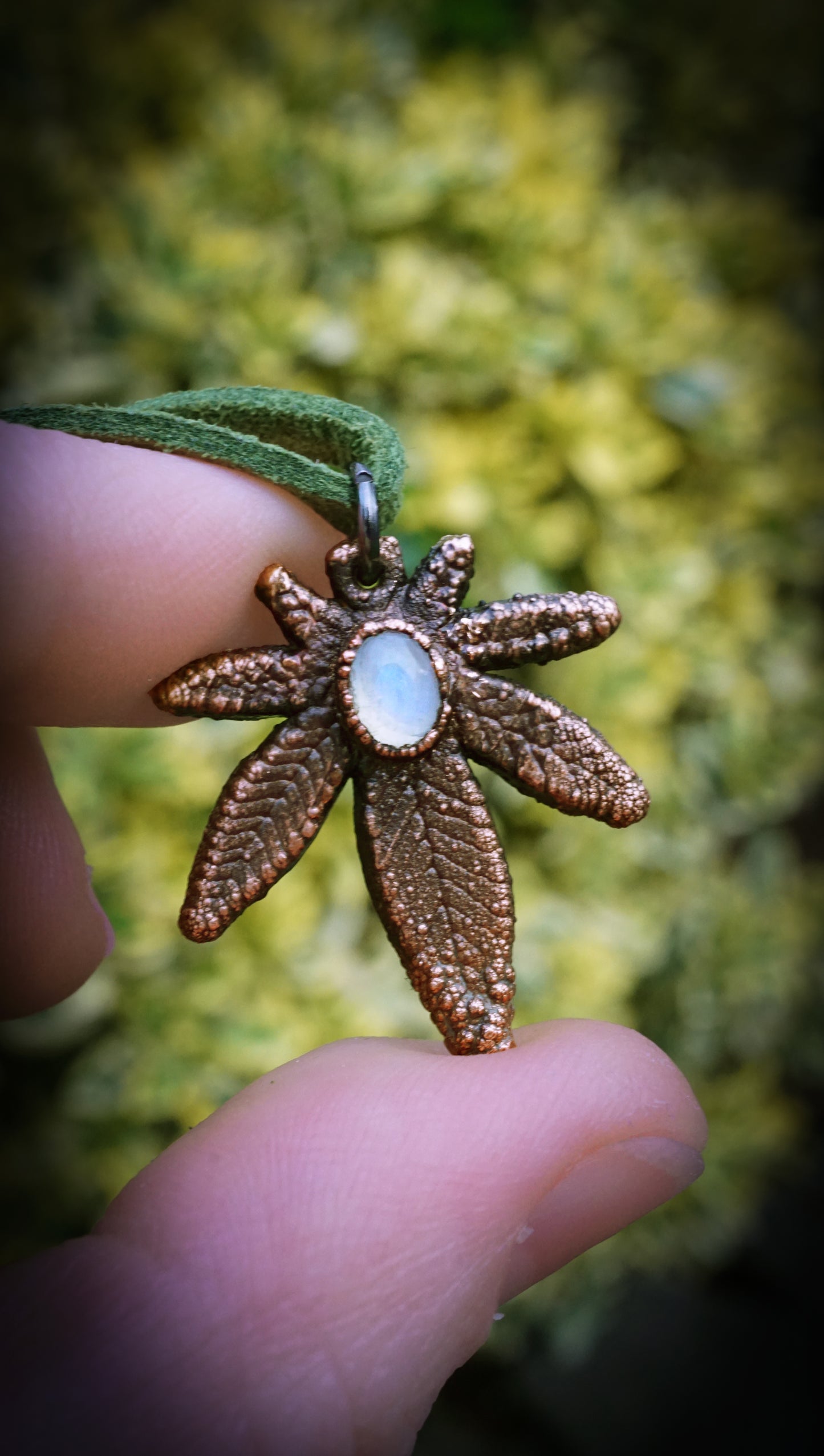 Bronze leaf-shaped pendant with a central gemstone held between fingers against a blurred natural background