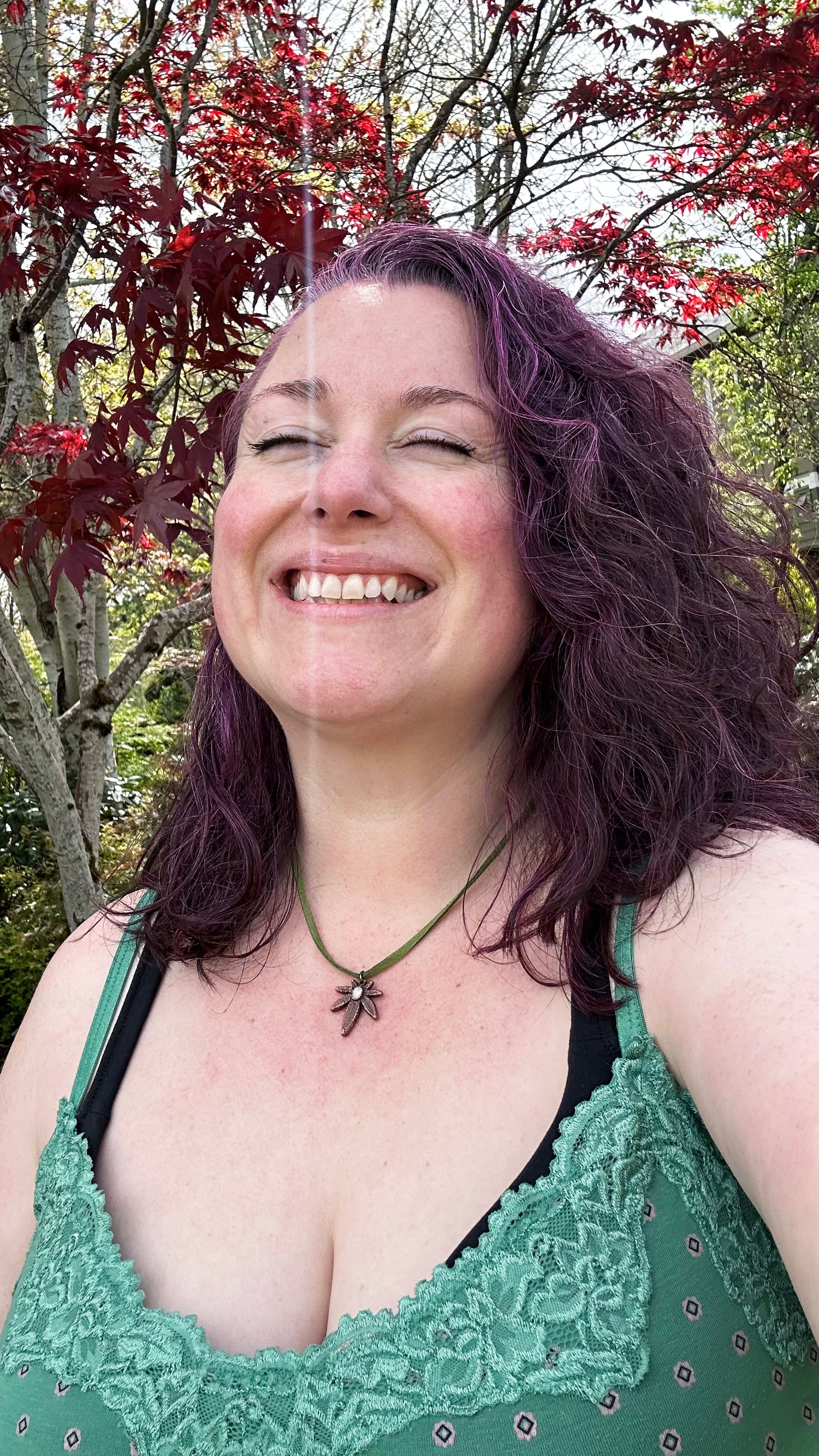 Woman with purple hair wearing a copper pot leaf necklace and green lace top, outdoors among trees with red leaves.