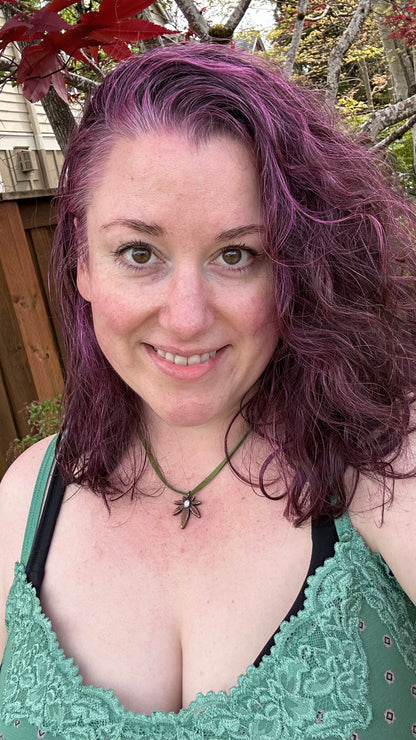 Woman with purple hair wearing a green lace top and copper leaf pendant outdoors.