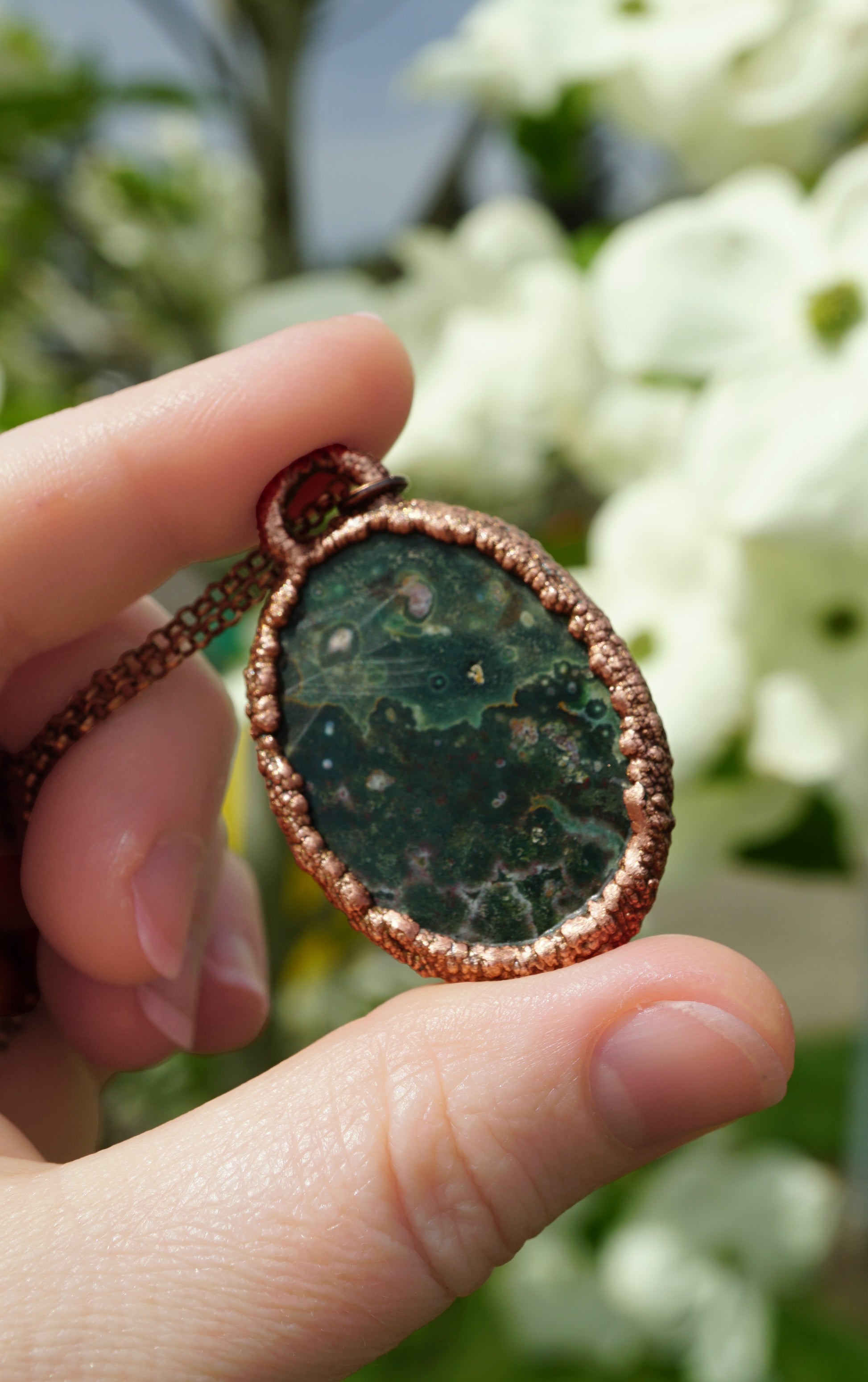 Hand holding a green stone pendant with a blurred floral background